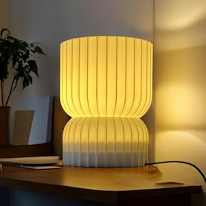 Front view of a modern, soft-glowing table lamp on a wooden table. The lamp provides a warm, diffused light, perfect for creating cozy ambient lighting in any living space.