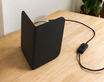 May include: A black table lamp with a simple design. The lamp has a square base and a single light bulb. The lamp is on a wooden table.
