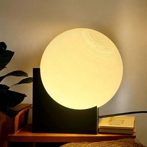 May include: A white globe-shaped lamp with a black square base sits on a wooden table. The lamp is turned on and emits a warm, white light.