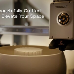 May include: A 3D printer with a white bowl and a white cylindrical object inside. The printer has a black nozzle and a white control panel with the text "Thoughtfully Crafted to Elevate Your Space".