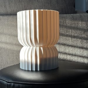 Modern table lamp, turned off, positioned on a black leather stool. Natural light highlights the lamp’s sculptural qualities, creating interesting shadows and emphasizing its elegant design.