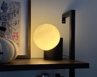 May include: A modern table lamp with a round, white, frosted glass shade and a black base. The lamp is on a wooden shelf.