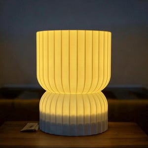 Front view of a minimalist table lamp on a wooden surface, casting a warm, ambient glow. The modern design and soft, diffused light enhance the cozy atmosphere of the space.