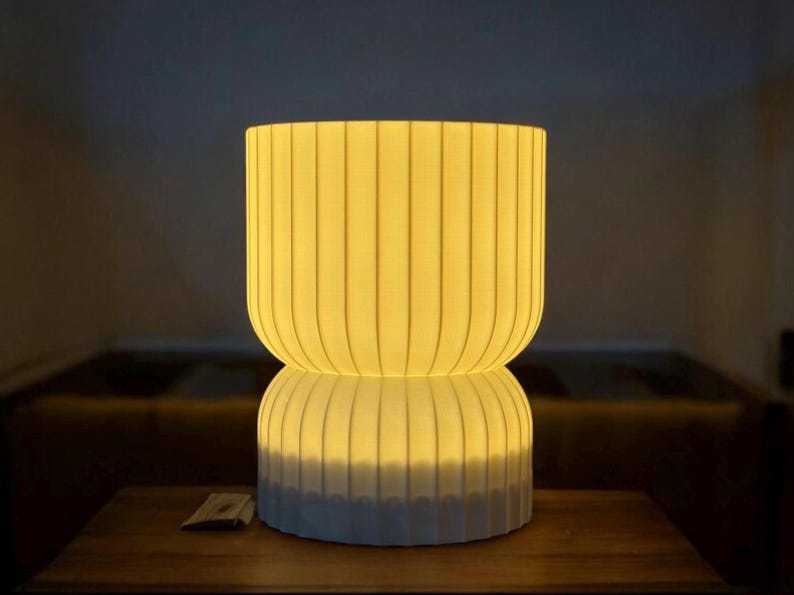 Front view of a minimalist table lamp on a wooden surface, casting a warm, ambient glow. The modern design and soft, diffused light enhance the cozy atmosphere of the space.