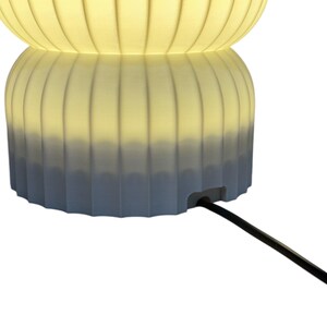 Close-up view of the bottom of a modern table lamp, turned on, showcasing the opening for the cord against a clean white background. The design emphasizes minimalist features and functionality.