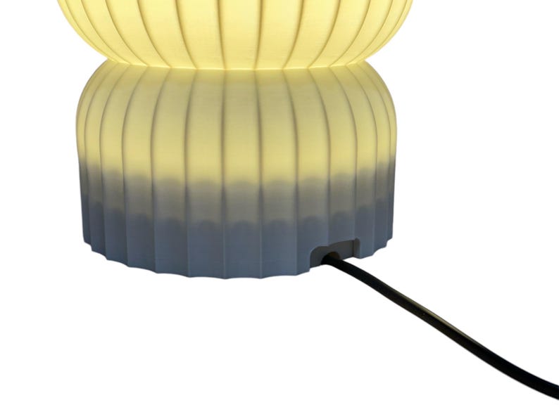 Close-up view of the bottom of a modern table lamp, turned on, showcasing the opening for the cord against a clean white background. The design emphasizes minimalist features and functionality.