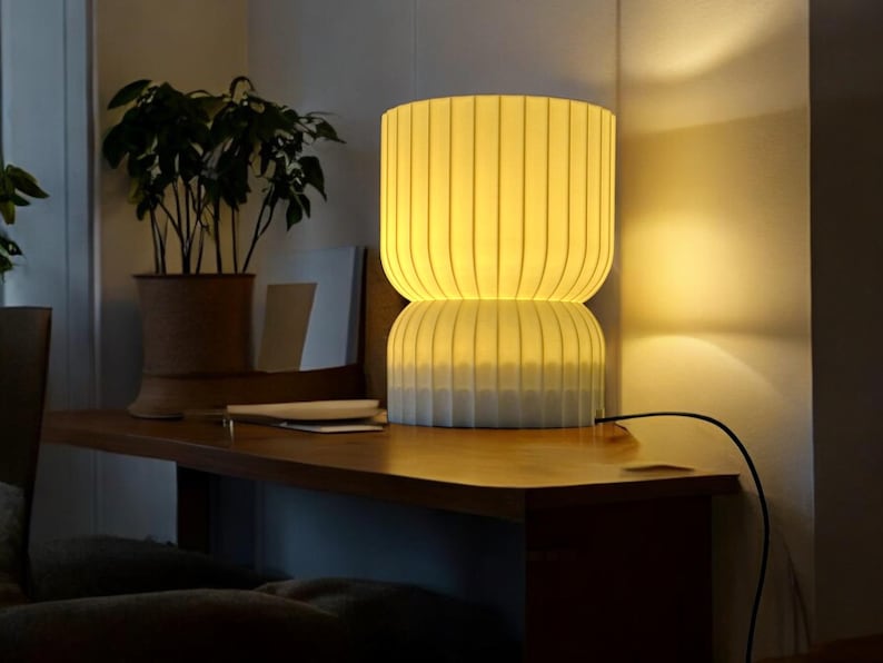 Front view of a modern, soft-glowing table lamp on a wooden table. The lamp provides a warm, diffused light, perfect for creating cozy ambient lighting in any living space.