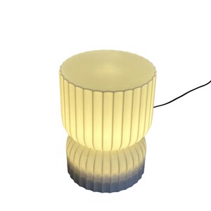 Full view of a modern table lamp, turned on, against a clean white background. Inspired by Japanese rice paper lamps, its minimalist design features soft lines, ready to emit a warm ambient glow when lit.