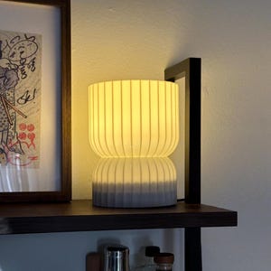 View of a modern table lamp emitting a soft, ambient glow, sitting on a sleek wooden shelf. The minimalist design complements the contemporary decor, adding warm lighting to the space.