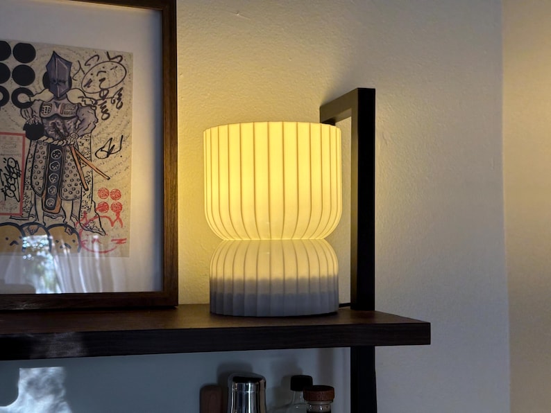 View of a modern table lamp emitting a soft, ambient glow, sitting on a sleek wooden shelf. The minimalist design complements the contemporary decor, adding warm lighting to the space.