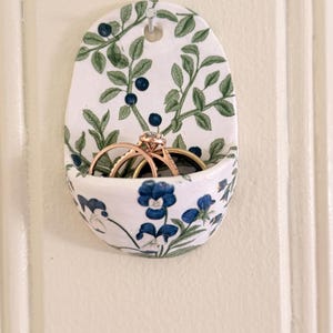 May include: A wall-mounted ceramic pocket holder featuring a white base with a botanical pattern of green leaves, dark blue berries, and blue flowers. The pocket contains two gold-toned rings, one of which is set with a clear gemstone.