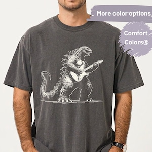 May include: Dark gray t-shirt featuring a white illustration of Godzilla playing a guitar. The shirt has a vintage, washed look. The text "Matty's T-Shirt Boutique" is in a brown frame. The shirt is part of the Comfort Colors® collection.