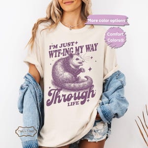 Op de afbeelding: Een cr&egrave;mekleurig t-shirt met een paarse opossum-afbeelding en de tekst "I'm just WTF-ing my way through life".
