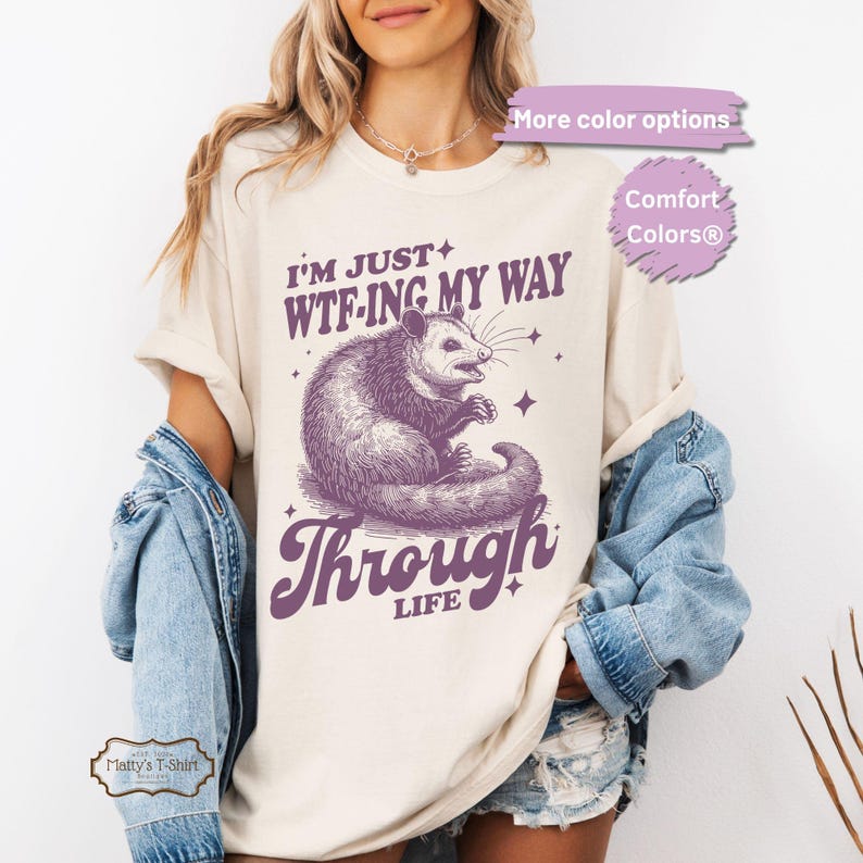 Op de afbeelding: Een cr&egrave;mekleurig t-shirt met een paarse opossum-afbeelding en de tekst "I'm just WTF-ing my way through life".