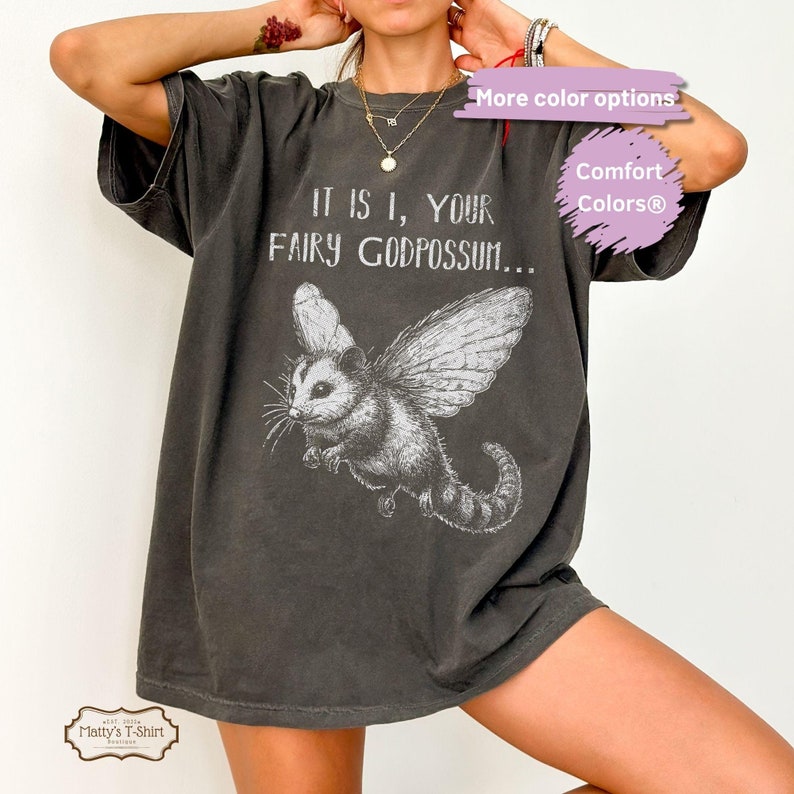 May include: A dark gray t-shirt with a graphic of a flying possum with wings and the text "IT IS I, YOUR FAIRY GODPOSSUM..." printed on it.