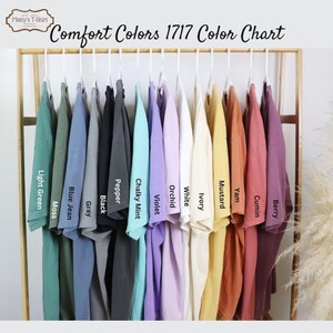 Op de afbeelding: Kleurenkaart met 14 verschillende kleuren T-shirts die aan een rek hangen. De kleuren zijn: Lichtgroen, Mos, Blauwe jeans, Grijs, Zwart, Peper, Krijtwitte munt, Violet, Orchidee, Wit, Ivoor, Mosterd, Yam, Komijn en Bes.