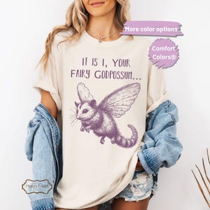 May include: A cream colored t-shirt with a purple illustration of a flying opossum with the text "IT IS I, YOUR FAIRY GODPOSSUM..." printed on the front.