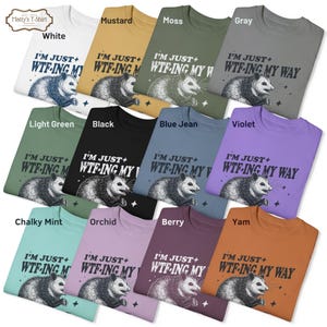Op de afbeelding: Een verzameling van 12 T-shirts met een opossum-afbeelding en de tekst "I'm just + WTF-ing my way". De shirts zijn in verschillende kleuren verkrijgbaar, waaronder wit, mosterd, mos, grijs, lichtgroen, zwart, jeansblauw, violet, krijtwit, orchidee, bes en yam.