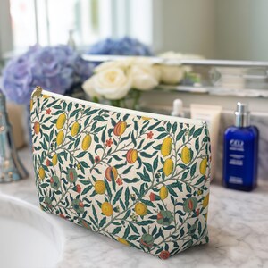 以下が含まれることがあります： レモン、桃、緑の葉が描かれた鮮やかな植物柄のクリーム色のコスメポーチ。ゴールドのジッパーが付いています。生地製と思われます。ヴィンテージのフローラルプリントを彷彿とさせるデザインです。