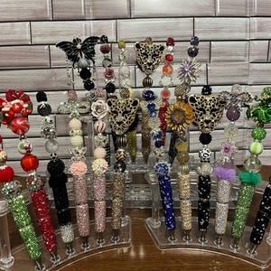 May include: A collection of decorative pens with various embellishments. The pens feature designs such as butterflies, leopard heads, flowers, and sunflowers. The pens are displayed on clear plastic stands, showcasing their colorful beads and sparkling details.