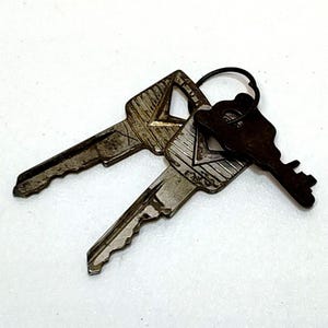 May include: Three metal keys on a white background. Two keys are silver-toned with a worn appearance, and the third is a darker, bronze color. All keys are attached to a black metal ring.