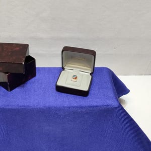 May include: A small, colorful pin with a logo inside a brown jewelry box. The pin features a design with red, blue, and yellow elements. The box is open, revealing the pin on a light gray interior. Another closed box is visible to the left.
