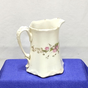 May include: White ceramic pitcher with a floral design in shades of pink and green. The pitcher has a handle and a scalloped rim.