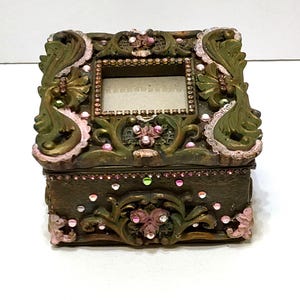 May include: Ornate, antique-style jewelry box in shades of green, pink, and gold. The box features a rectangular photo frame on the lid, embellished with rhinestones and floral accents. The box is intricately carved with scrollwork and decorative elements.