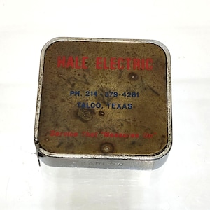 May include: Vintage Hale Electric tape measure with a brass-colored top and a silver-colored metal casing. The top features red text that reads "HALE ELECTRIC" and blue text with a phone number and location.