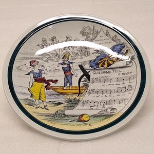 May include: A decorative ceramic plate with a detailed illustration featuring figures in period clothing, a boat, and musical notes. The plate has a white rim with a green and blue border, and the text "Guillaume Tell" is visible.