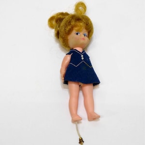 May include: A small plastic doll with blonde hair wearing a blue dress. The doll has blue eyes and a pink face. The doll is standing on a white surface.