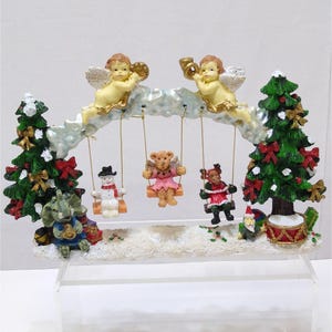 May include: A Christmas-themed figurine with two angels on a cloud swing set.  The swing set has four figures on swings: a snowman, a teddy bear in a pink dress, a reindeer in a red dress, and a black and white figure.  The scene is set in a snowy forest with two Christmas trees.