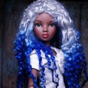 May include: A doll with long, curly, ombre hair that transitions from white to blue. The doll is wearing a white shirt and blue jeans.