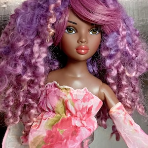 May include: A fashion doll with dark skin, wearing a pink floral top and denim shorts. The doll has large green eyes, full lips, and long, curly purple and pink hair. The doll's top has a ruffled neckline and long, sheer sleeves.