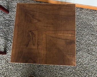 May include: Square wooden side table with a dark brown finish. The tabletop features a chevron pattern. The table is approximately 24 inches wide and 24 inches long.