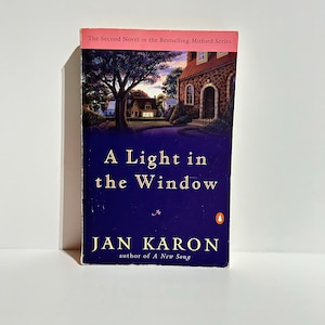 May include: A hardcover book titled "A Light in the Window" by Jan Karon. The cover features a nighttime landscape with a house and a tree. The book is part of the Bestselling Mitford Series.