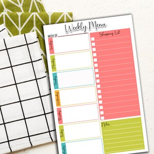 May include: A weekly menu and shopping list notepad with a white background. The notepad has a pink shopping list section and a green notes section. The days of the week are in different colors. Two patterned kitchen towels are in the background.