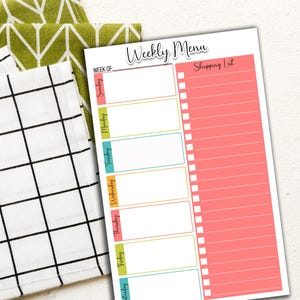 May include: A weekly menu and shopping list planner with a white background. The planner has a coral pink shopping list section and a section for each day of the week. The words "Weekly Menu" are at the top. Two decorative kitchen towels are in the background.