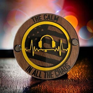 May include: Round wooden coaster with the text "THE CALM IN ALL THE CHAOS". The coaster features a yellow design of headphones and a heartbeat over a black and white American flag.
