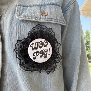 May include: A round white pin-back button featuring the black text WOO PIG! in a bold, retro-style font. The button is framed by a ruffled layer of black lace trim, creating a decorative circular accessory attached to a light blue denim jacket.