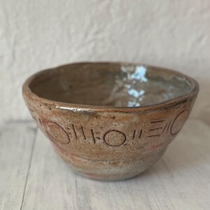 May include: A handmade ceramic bowl with a rustic, earthy aesthetic. The bowl features a light brown exterior with etched geometric patterns and subtle red accents. The interior has a glazed finish.