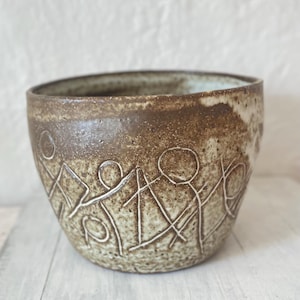 May include: A handcrafted, brown ceramic bowl with a textured surface. The bowl features a white, etched design of stick figures holding hands around the exterior. The interior has a light blue glaze. The bowl is a decorative piece.