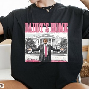 May include: Black t-shirt with the text "DADDY'S HOME" in pink. The graphic features a man in a suit with arms outstretched in front of the White House. The word "BLACK" is at the bottom of the shirt.