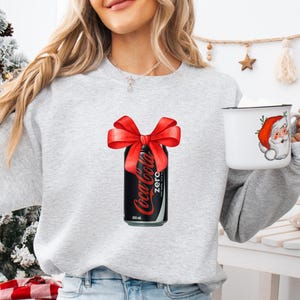 May include: A gray sweatshirt with a Coca-Cola Zero can with a red bow on top. The can is 330 ml.