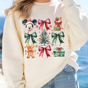 May include: A cream-colored sweatshirt with a Christmas-themed design featuring Mickey Mouse, a gingerbread man, a Christmas tree, and presents. The design is in watercolor style with red, green, and brown accents.