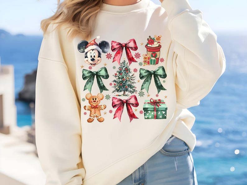 May include: A cream-colored sweatshirt with a Christmas-themed design featuring Mickey Mouse, a gingerbread man, a Christmas tree, and presents. The design is in watercolor style with red, green, and brown accents.