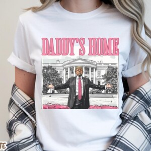 May include: White t-shirt with the text "DADDY'S HOME" in pink letters above an illustration of a man in a suit with his arms outstretched in front of the White House.