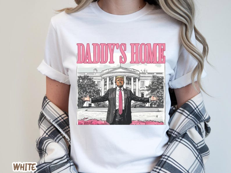 May include: White t-shirt with the text "DADDY'S HOME" in pink letters above an illustration of a man in a suit with his arms outstretched in front of the White House.