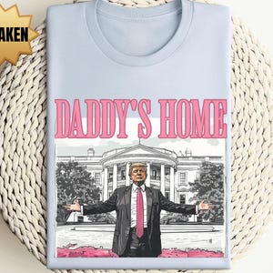 May include: Light blue t-shirt with the text "DADDY'S HOME" in pink. The graphic features a man in a suit with the White House in the background. The word "UNSHAKEN" is in a gold starburst.