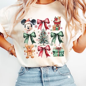May include: A white t-shirt with a Christmas-themed design featuring Mickey Mouse, a gingerbread man, a Christmas tree, presents, and bows. The design is in a watercolor style with red, green, and brown colors.